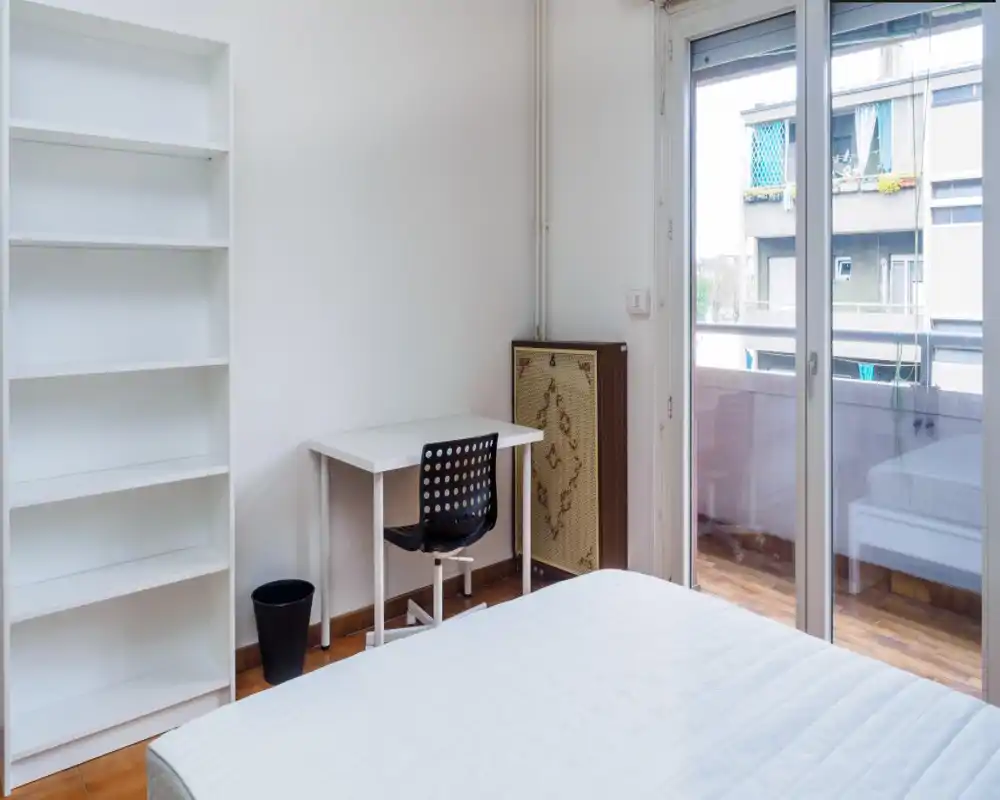 Dormitorio luminoso y minimalista con cama blanca hecha, un escritorio y silla junto a la puerta del balcón que aporta abundante luz natural; estantería y suelo de parquet completan el espacio ordenado.