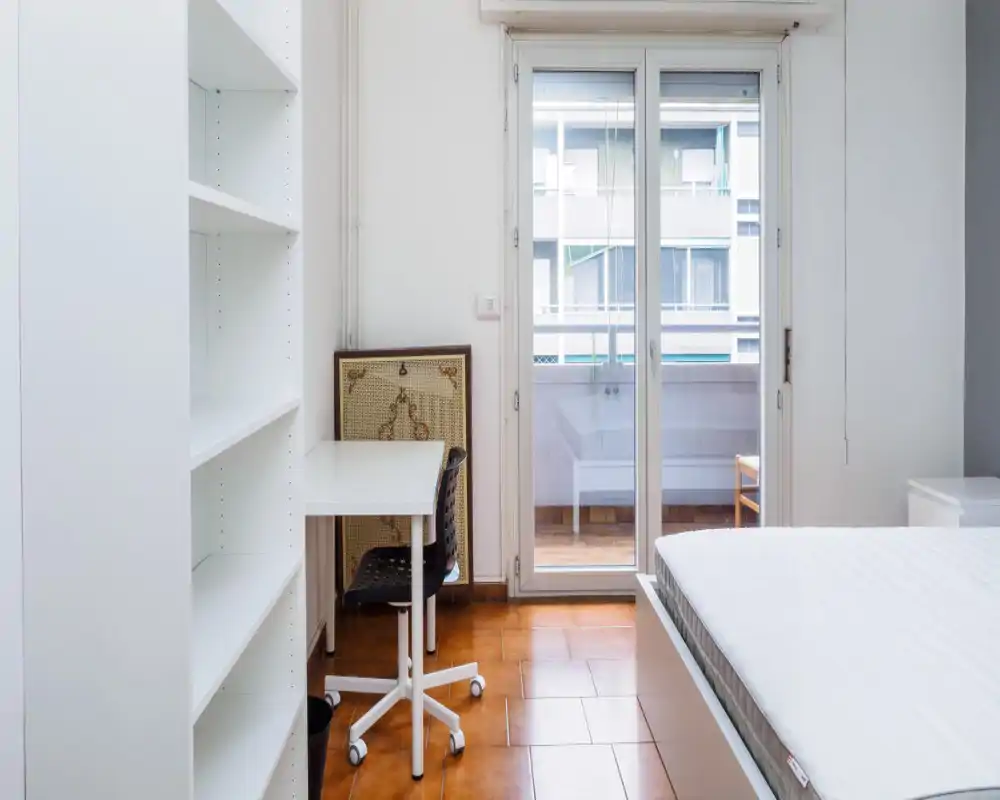 Dormitorio luminoso y minimalista con cama doble visible, escritorio y estantería junto a la puerta del balcón que aporta luz natural—apelo limpio y moderno para anuncios.