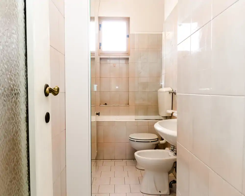 Baño compacto y limpio con bañera, inodoro, bidé y lavabo. Baldosas neutras y luz natural de una pequeña ventana crean un espacio simple y funcional.