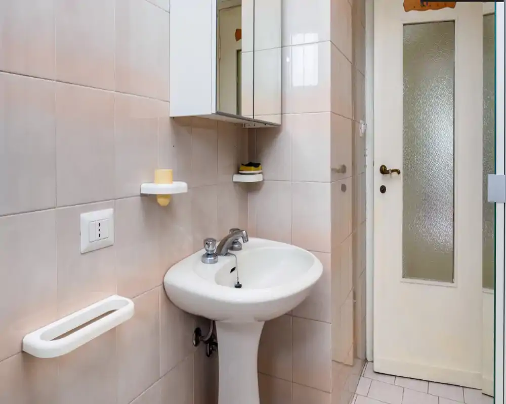 Baño compacto y bien iluminado con lavabo de pedestal, armario con espejo y paredes alicatadas; distribución funcional y limpia, apropiado para un anuncio.