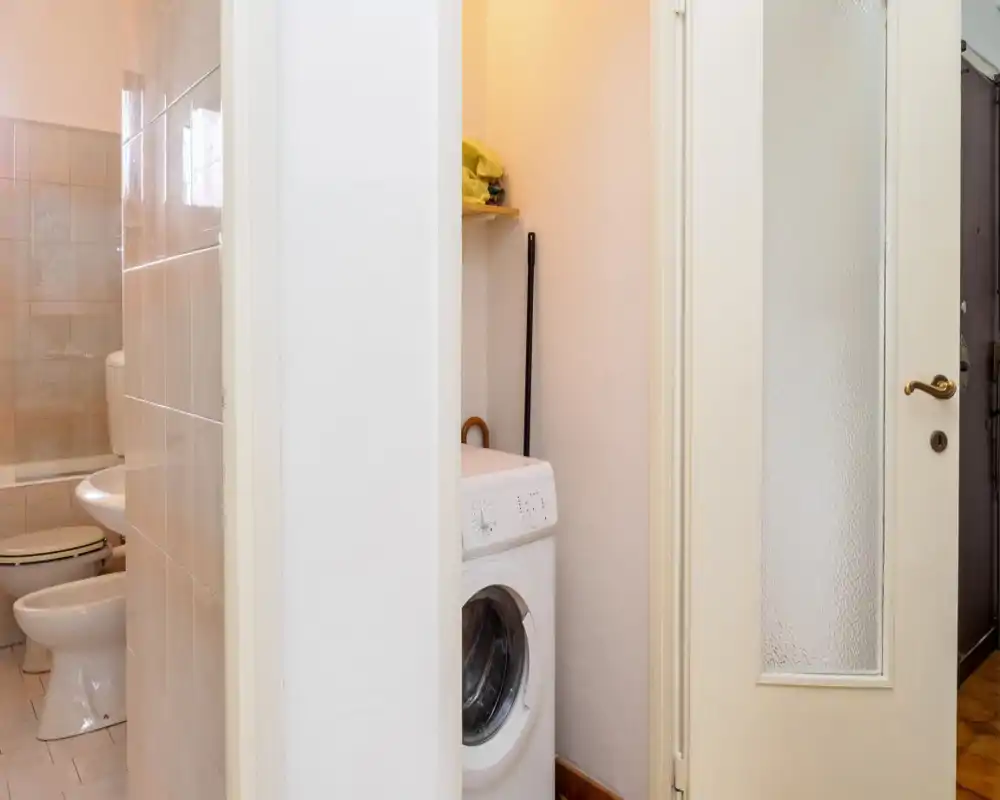 Baño compacto con inodoro, bidé y zona de ducha visibles; una lavadora se encuentra en un pequeño hueco contiguo. Buena iluminación y revestimiento cerámico limpio, espacio funcional.