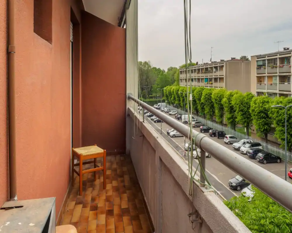 Balcón estrecho con suelo tipo terracota, un sencillo taburete de madera y barandilla metálica con vista a una calle arbolada y coches aparcados — ideal para mostrar el espacio exterior y las vistas.