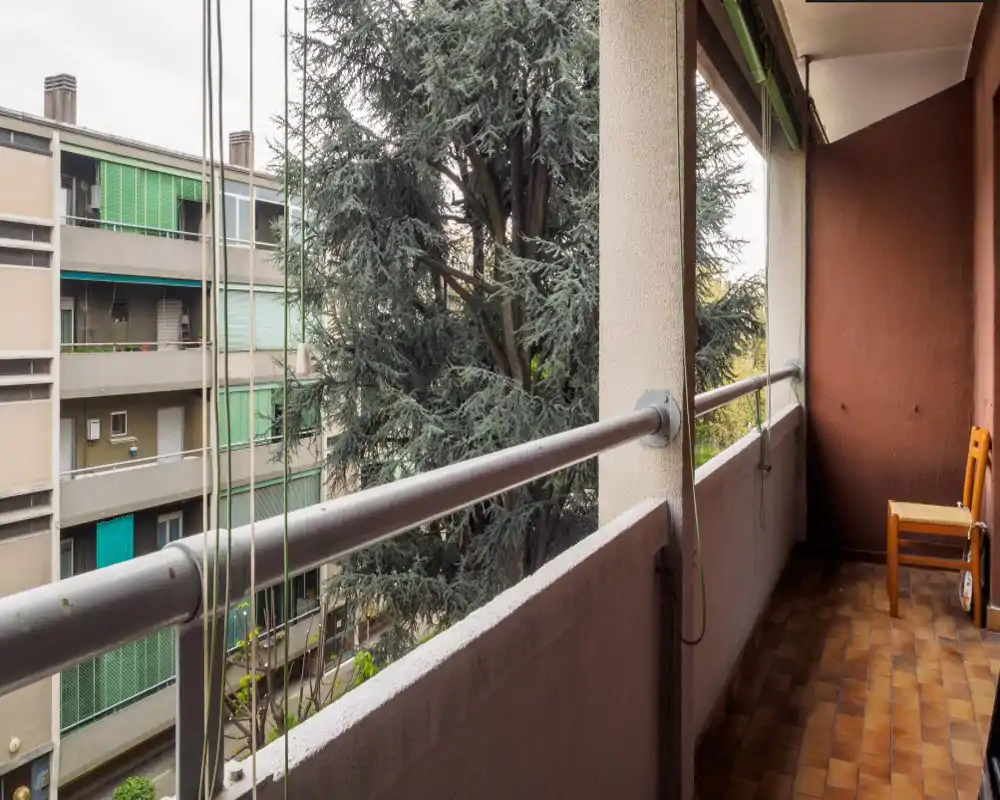 Balcón cubierto con suelo de baldosas y barandilla, ideal para sillas o plantas; vistas a edificios y árboles cercanos.