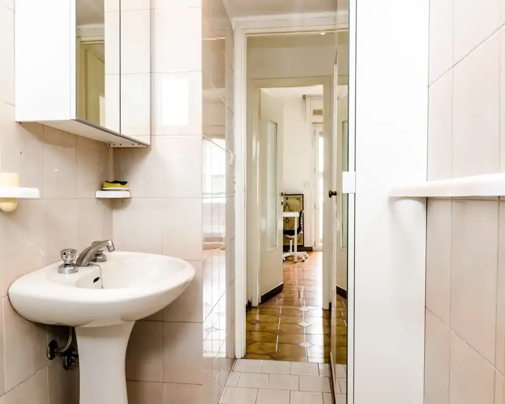 Baño luminoso y limpio con lavabo sobre pedestal, armario con espejo montado en la pared y azulejos; la puerta da a un pasillo con suelo de madera.