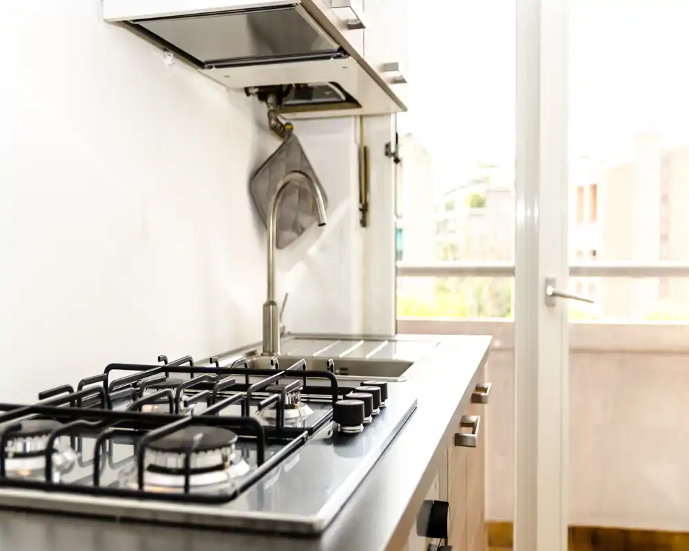 Rincón de cocina moderno y luminoso con encimera de gas, fregadero de acero inoxidable y una gran puerta de cristal que aporta luz natural — limpio y funcional.