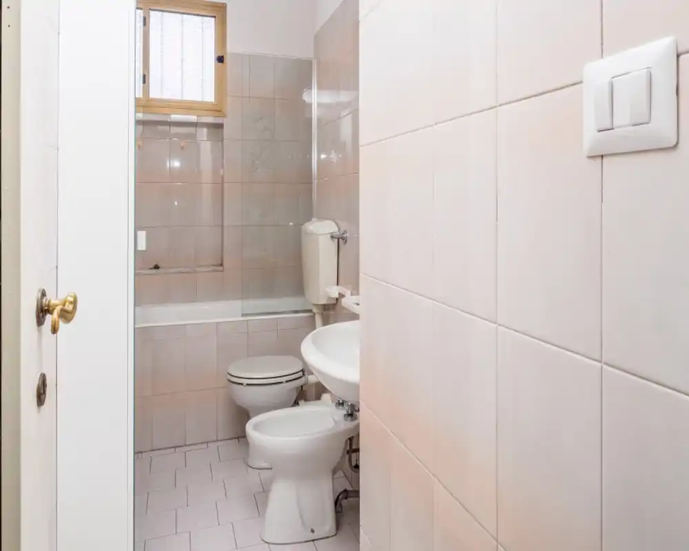 Baño pequeño alicatado con bañera, inodoro y bidé; azulejos claros y luz natural de una ventana pequeña que crean un espacio limpio y funcional.