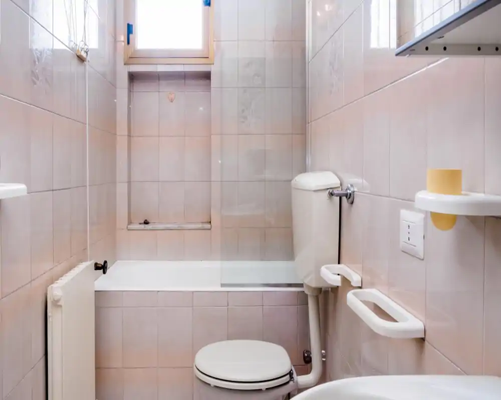 Baño compacto alicatado con bañera, inodoro y accesorios de pared, iluminado por una pequeña ventana que aporta luz natural suave.