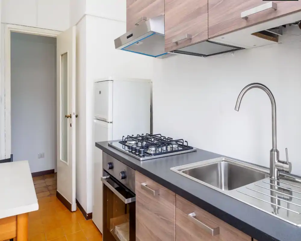 Cocina moderna y luminosa con placa de gas, horno empotrado, fregadero de acero inoxidable y muebles efecto madera — espacio de cocinado compacto y ordenado ideal para un piso pequeño.