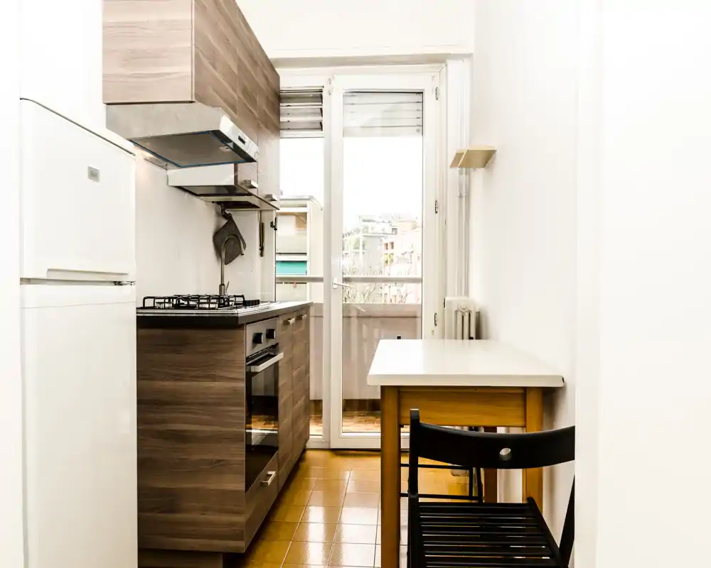 Cocina compacta y moderna con muebles efecto madera, placa de gas, horno empotrado y una pequeña mesa junto a la puerta acristalada que da al balcón — luminosa y bien organizada para vivir en la ciudad.