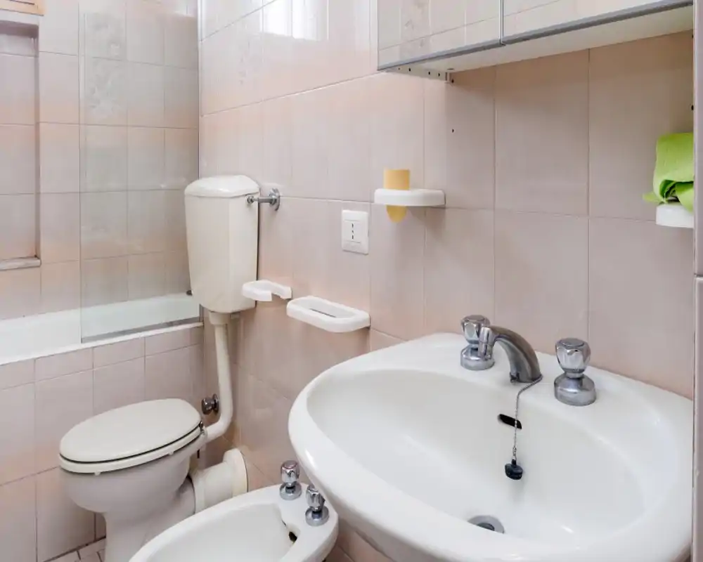Baño pequeño con azulejos que incluye lavabo, bidé, inodoro y bañera parcialmente visible; azulejos claros y grifería cromada que transmiten un aspecto limpio y funcional.