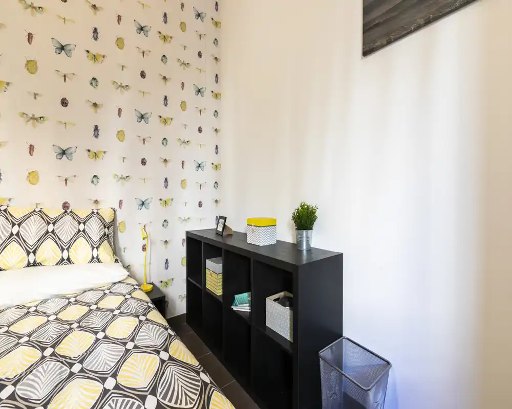 Rincón de dormitorio luminoso y estiloso con ropa de cama estampada y papel pintado de mariposas; estantería negra con cestas y planta que aporta orden y encanto.