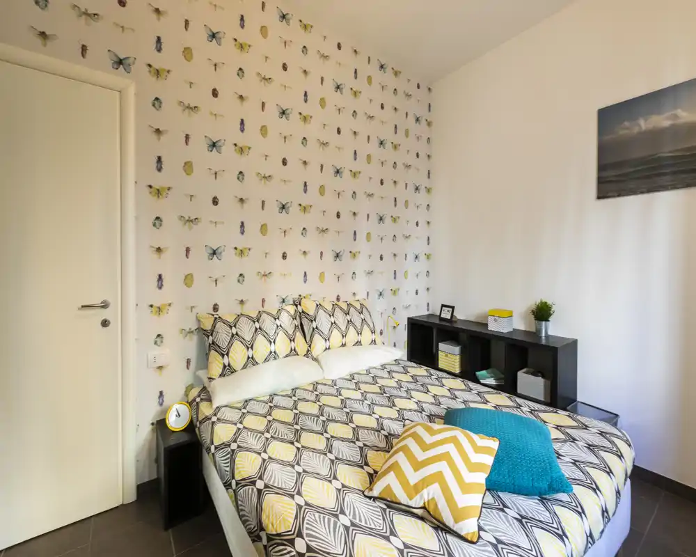 Dormitorio luminoso y moderno con ropa de cama estampada y papel pintado de mariposas; toques amarillos y estanterías que aportan calidez.