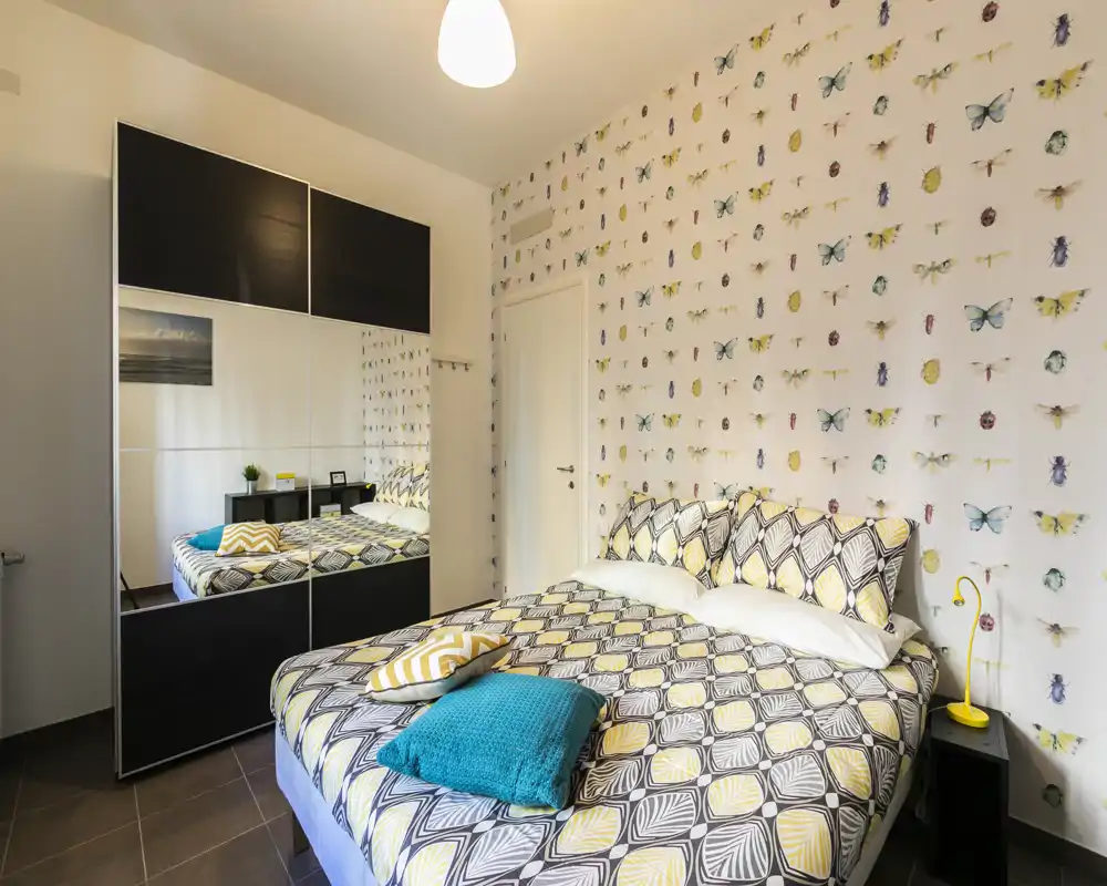 Dormitorio luminoso y moderno con colcha estampada, cojines coloridos y armario con espejo que amplía el espacio. Pared decorativa con mariposas y lámpara de mesita que aportan calidez.