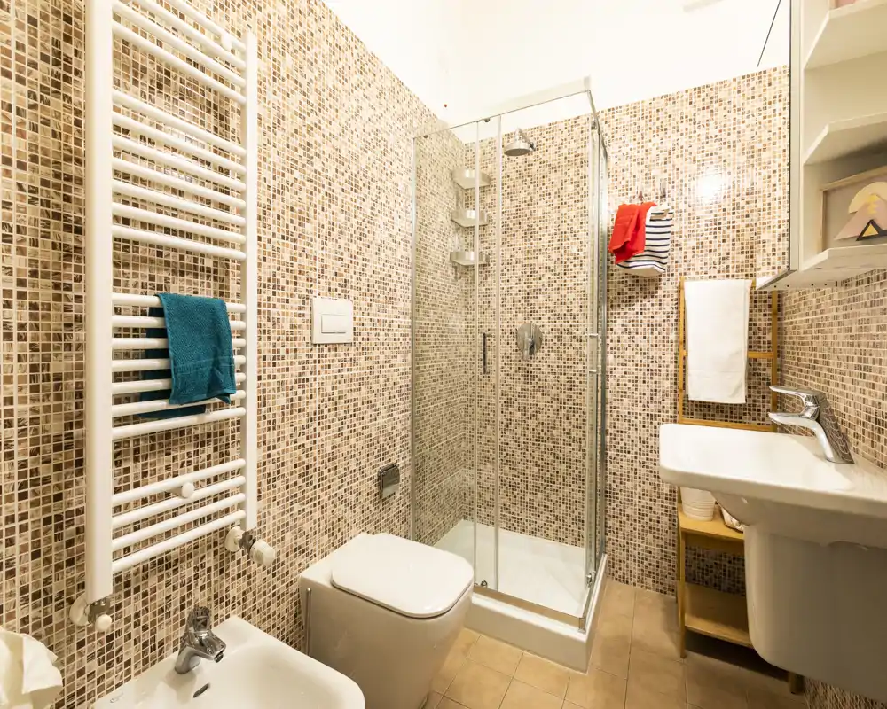 Baño moderno y luminoso con azulejos de mosaico, ducha acristalada, inodoro suspendido y lavabo de pedestal — compacto y bien iluminado.