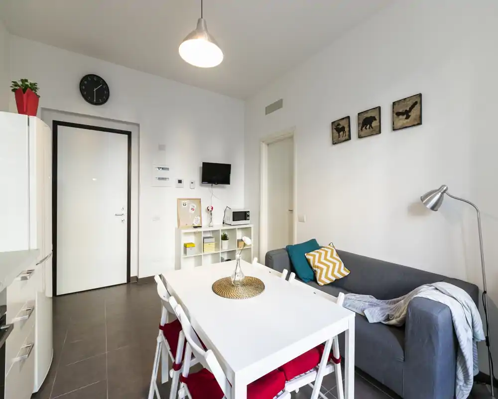 Zona de estar moderna y luminosa con mesa de comedor y sofá cómodo. El espacio presenta líneas limpias, cojines coloridos y estanterías prácticas — ideal para un anuncio de piso compacto.