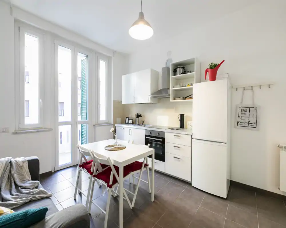 Zona de día moderna y luminosa con cocina compacta blanca, mesa de comedor y un pequeño sofá — ideal para un piso en la ciudad. Grandes ventanas que proporcionan abundante luz natural y un aspecto contemporáneo y limpio.