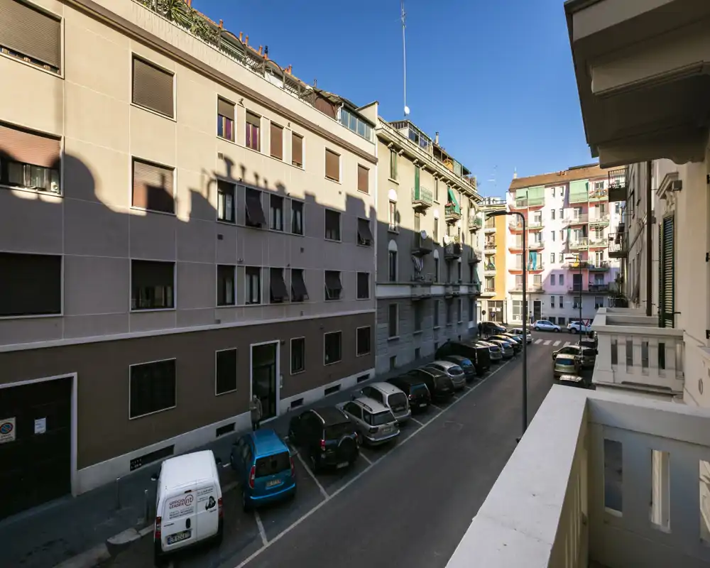 Vista soleada de la calle desde un balcón privado con coches aparcados y fachadas de edificios cercanos — cielo azul y buena luz natural.