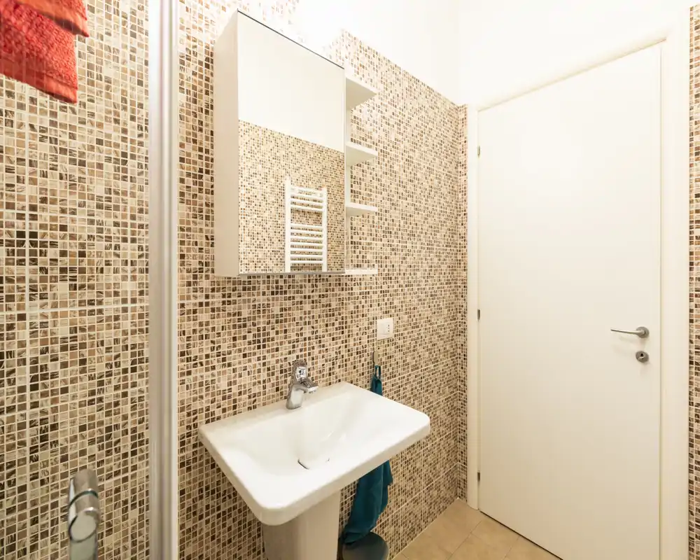 Baño moderno y luminoso con paredes de mosaico, armario con espejo y lavabo sobre pedestal — limpio y listo para huéspedes.