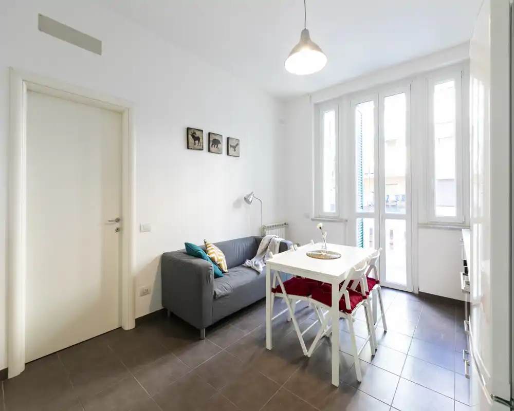 Área de salón-comedor luminosa y moderna con un sofá gris compacto, mesa de comedor blanca y grandes ventanas que aportan abundante luz natural — ideal para un apartamento urbano acogedor.