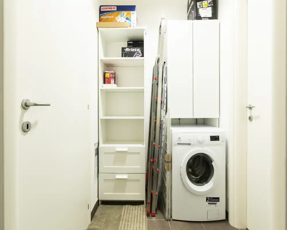 Área de lavandería/servicio compacta y luminosa con lavadora de carga frontal, estanterías altas y armarios: almacenamiento funcional para un piso pequeño.