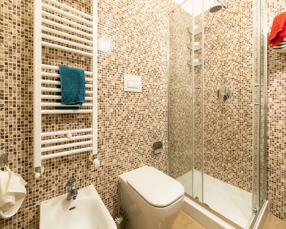 Baño moderno y compacto con paredes de mosaico, mampara de ducha de cristal, inodoro suspendido y bidé; iluminación cálida y accesorios contemporáneos.
