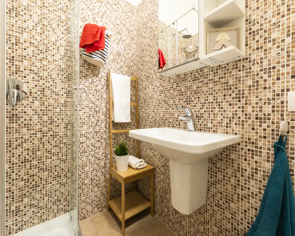 Baño moderno y luminoso con paredes de mosaico, lavabo mural, mampara de ducha de cristal y toallas y accesorios coordinados.