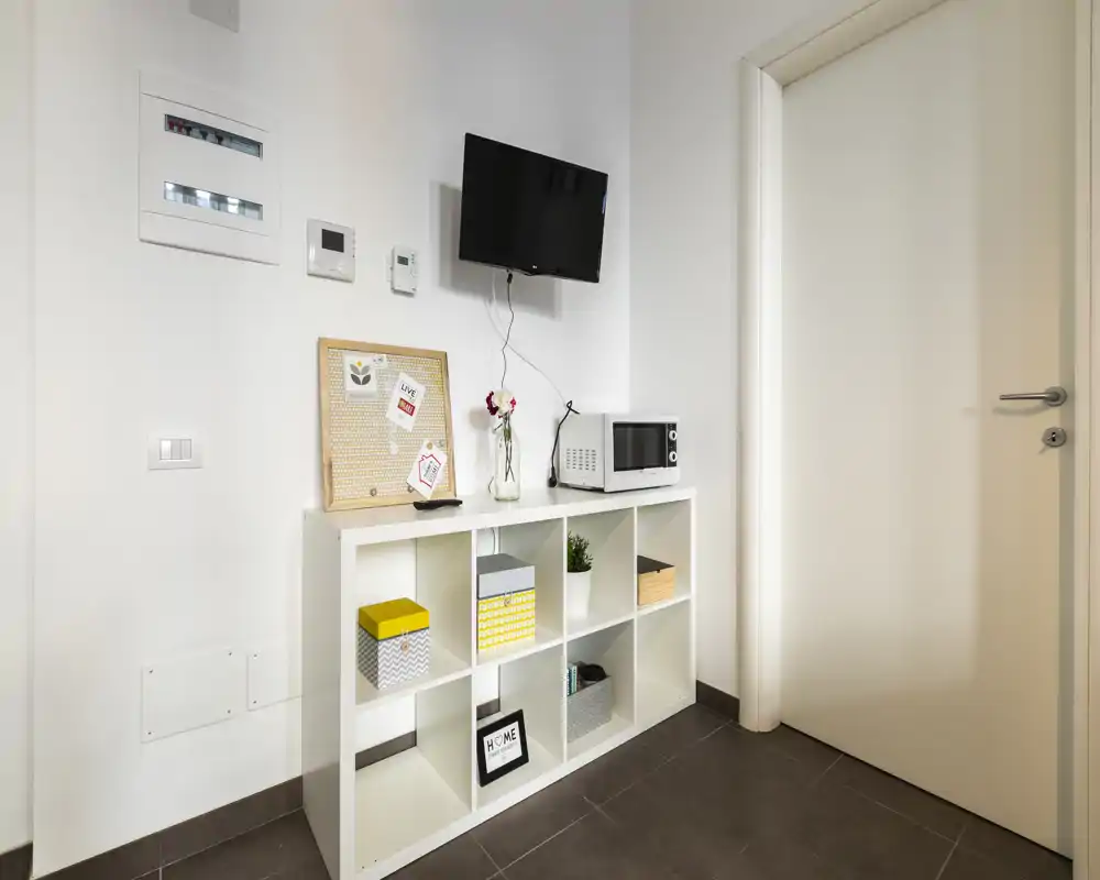 Rincón de entrada luminoso y minimalista con estantería de cubos, microondas pequeño y televisor en la pared — decoración limpia y neutra perfecta para la zona de entrada.