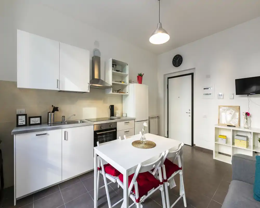 Cocina abierta moderna con armarios blancos, electrodomésticos de acero y mesa central con cojines rojos — luminosa, limpia e ideal para comidas en pequeño grupo.