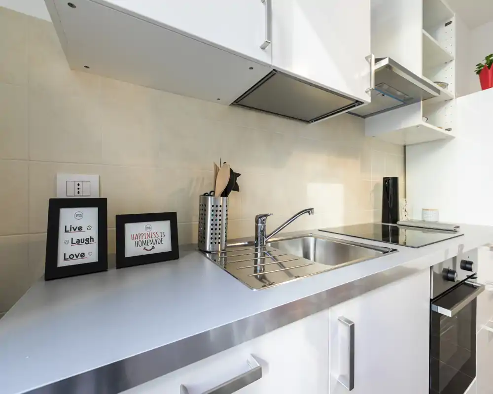 Cocina moderna y compacta con fregadero de acero inoxidable, placa de inducción y muebles blancos; decorada con marcos y utensilios que la hacen acogedora.