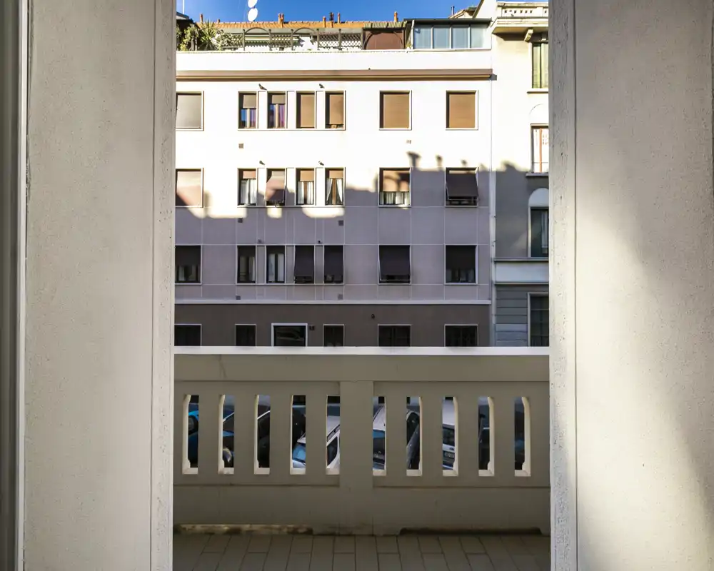 Pequeño balcón urbano con barandilla, vista a la fachada de un edificio contiguo y coches aparcados. Iluminación diurna y buena nitidez que muestran claramente el espacio exterior.