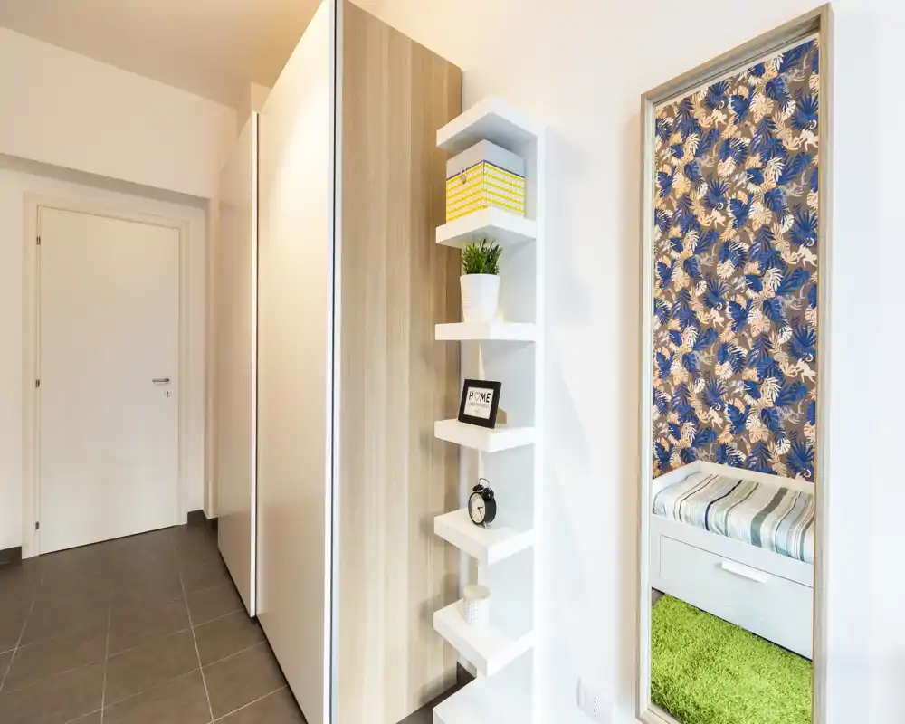 Recibidor luminoso y moderno con armario empotrado, estanterías decorativas y un espejo de cuerpo entero que refleja un pequeño rincón con asiento.