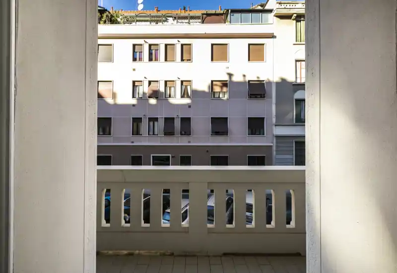 Pequeño balcón urbano con barandilla, vista a la fachada de un edificio contiguo y coches aparcados. Iluminación diurna y buena nitidez que muestran claramente el espacio exterior.