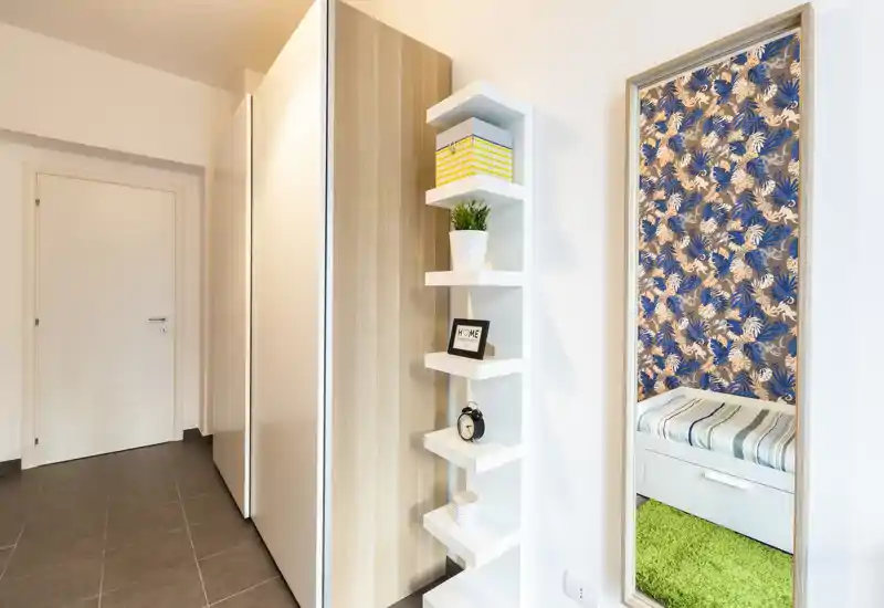 Recibidor luminoso y moderno con armario empotrado, estanterías decorativas y un espejo de cuerpo entero que refleja un pequeño rincón con asiento.