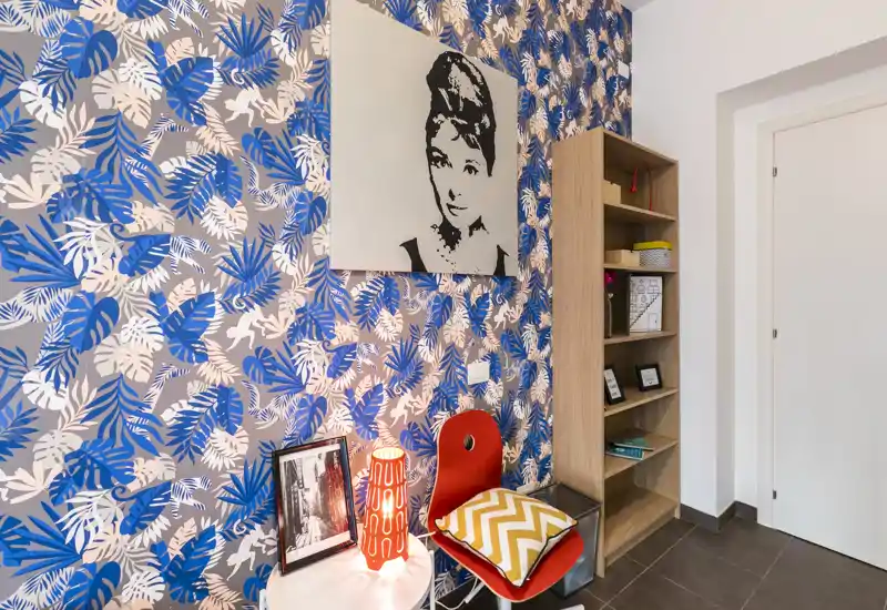 Rincón de estar colorido con papel pintado botánico, silla roja con cojín, mesita y estantería — ideal como zona de lectura o asiento de entrada.