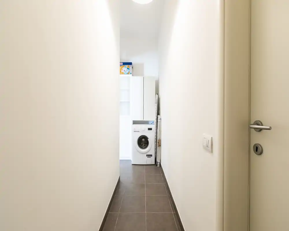 Entrada/pasillo estrecho con suelo de baldosas y lavadora visible al fondo; paredes blancas limpias y acabados modernos sencillos.