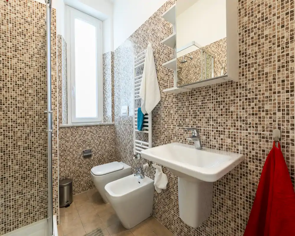 Baño moderno y compacto con paredes de mosaico, lavabo suspendido, bidé y inodoro, toallero calefactado y ventana translúcida que aporta luz natural.