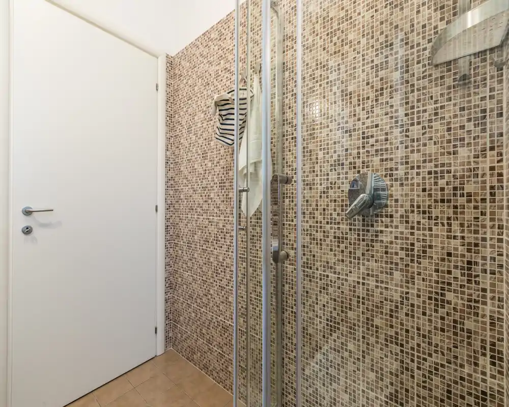 Baño compacto con ducha acristalada y alicatado de mosaico marrón con grifería cromada; espacio sencillo y bien iluminado.