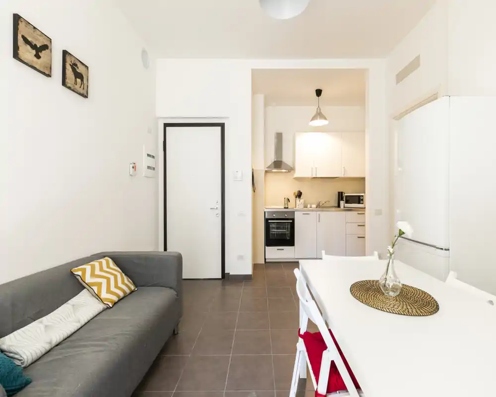 Zona de día luminosa y moderna en concepto abierto con sofá gris, mesa de comedor y una cocina blanca compacta — ideal para un piso pequeño o estudio.