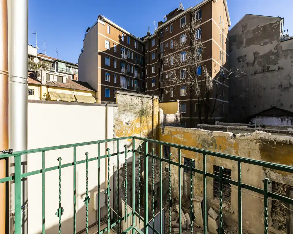 Pequeño balcón soleado con barandilla metálica verde que da a un patio interior y a los edificios circundantes.