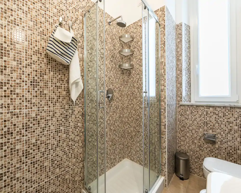Baño moderno con ducha acristalada, paredes de mosaico y inodoro blanco junto a una ventana esmerilada — limpio y luminoso.