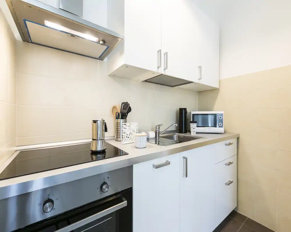 Cocina moderna y compacta con armarios blancos, electrodomésticos de acero inoxidable y placa de inducción. Salpicadero de azulejos, fregadero integrado y pequeños electrodomésticos para una cocina eficiente.