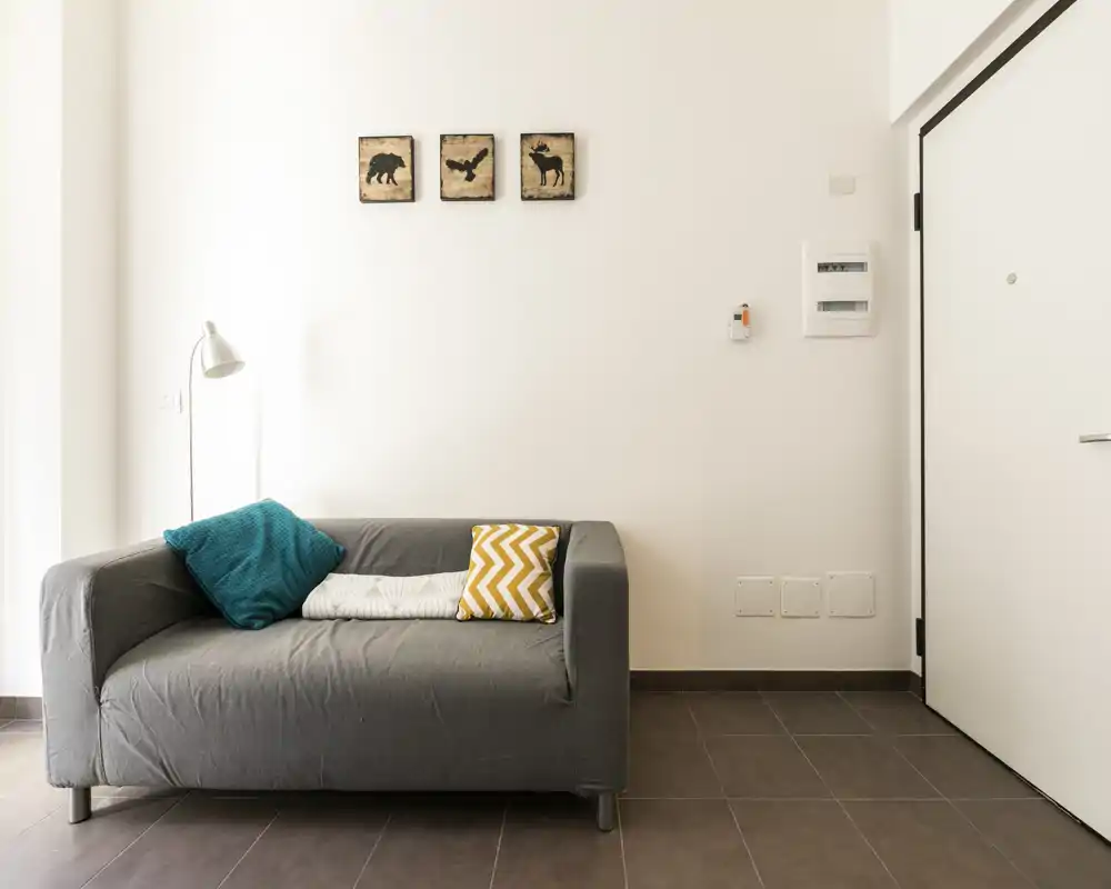 Área de estar minimalista con un sofá gris de dos plazas, cojines coloridos y una lámpara de pie frente a una pared blanca luminosa — un rincón lounge compacto y moderno.