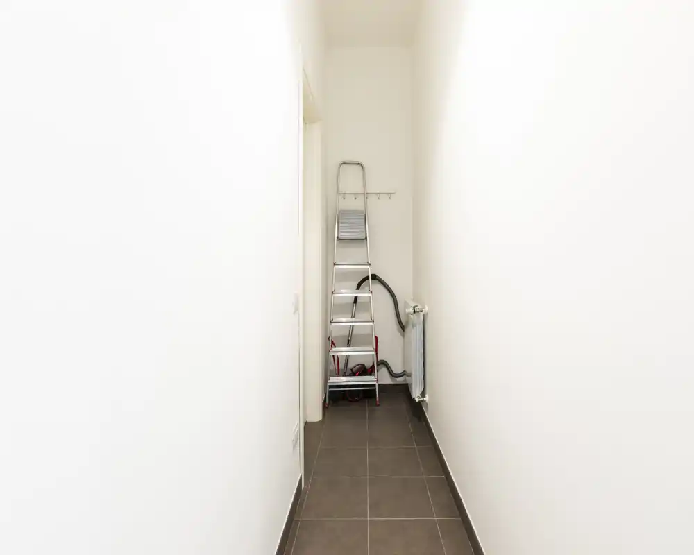 Entrada/pasillo estrecho con suelo de baldosas, paredes blancas y una escalera plegable al fondo: un paso sencillo y funcional.