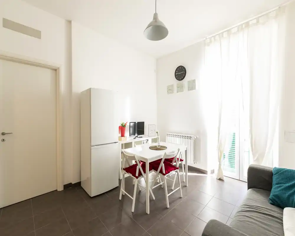 Espacio de día moderno y luminoso con mesa de comedor, frigorífico y sofá cómodo — ideal para un apartamento compacto. Gran ventana con cortinas ligeras que llena la estancia de luz natural.