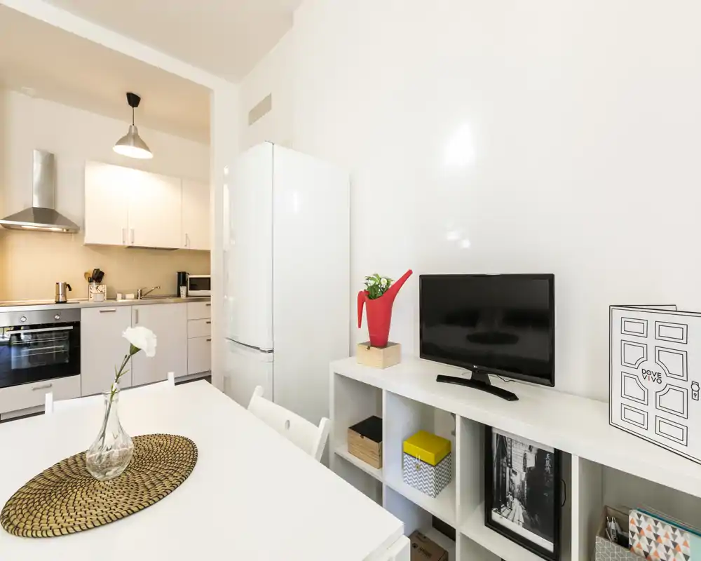 Cocina abierta moderna y luminosa con mesa de comedor, muebles blancos, frigorífico y estantería compacta — ideal para un apartamento pequeño.