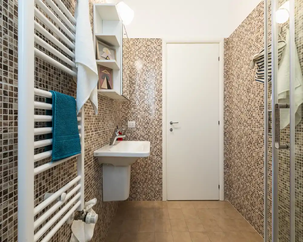 Baño compacto y moderno con paredes de mosaico, lavabo de pared, radiador toallero y mampara de ducha de cristal. Espacio luminoso, limpio y bien presentado para un anuncio.