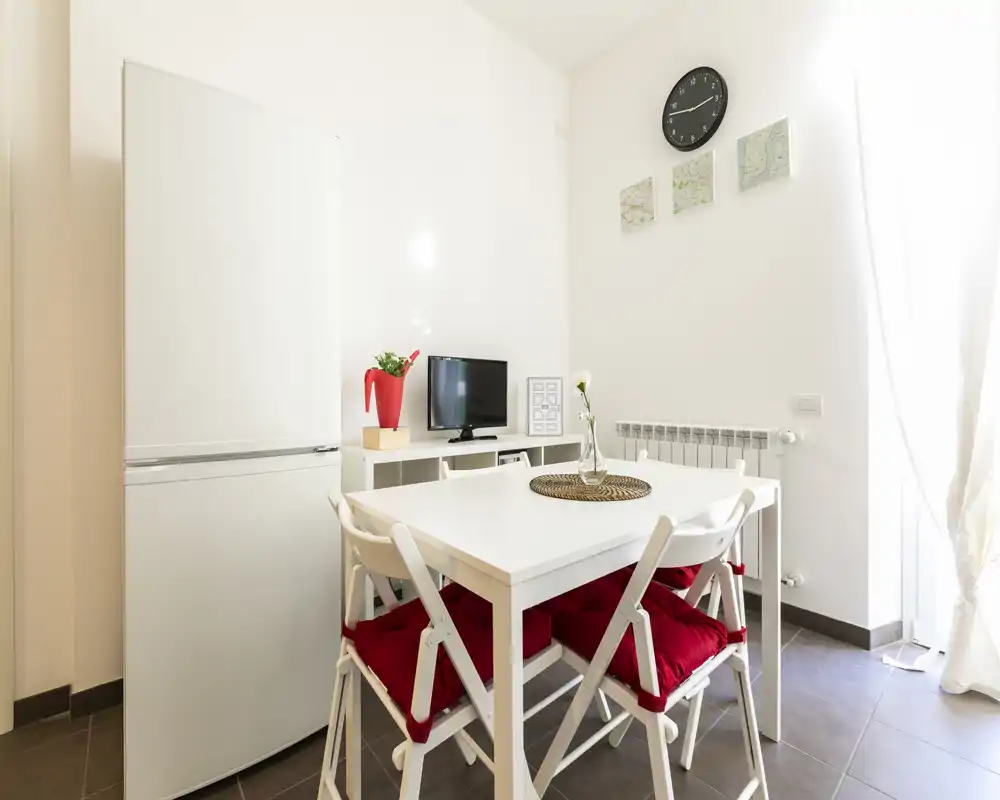Área de comedor compacta y luminosa con mesa blanca y cuatro sillas con cojines rojos, pequeña consola de TV y frigorífico visibles, ideal para un apartamento pequeño.
