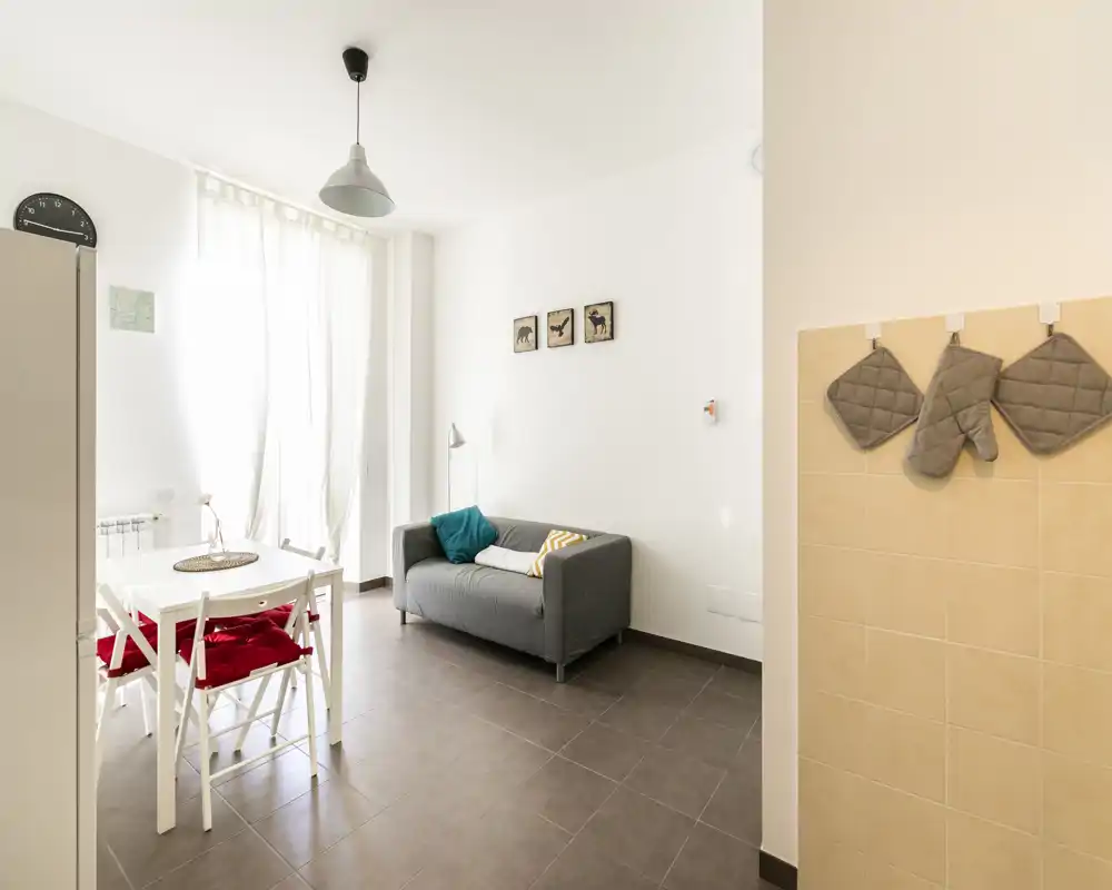 Sala luminosa y minimalista con un pequeño sofá gris, mesa de comedor con sillas con cojines rojos y una gran ventana que deja entrar luz natural — ideal para un apartamento compacto.