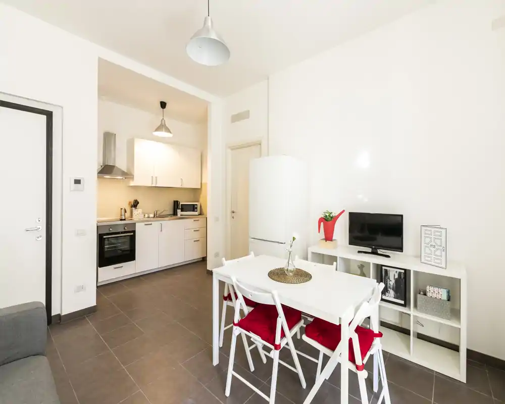 Zona de estar abierta y moderna con cocina compacta, mesa blanca con sillas de cojines rojos y pequeño mueble TV — ideal para un piso en la ciudad.