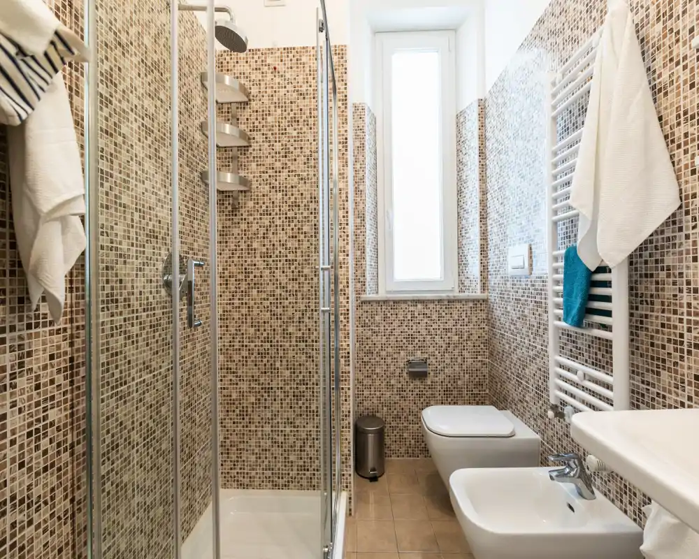 Baño moderno y luminoso con ducha acristalada, bidé, inodoro y radiador toallero mural; paredes en mosaico y una ventana estrecha que aporta luz natural.